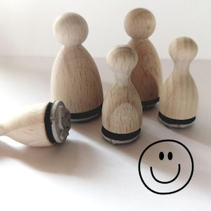 Könnte beinhalten: Eine Sammlung von fünf hellen Holzstempeln mit abgerundeten Oberseiten und flachen Unterseiten. Vier Stempel stehen aufrecht, während einer auf der Seite liegt und eine graue Gummistempelfläche zeigt. Eine einfache schwarze Strichzeichnung eines Smileys ist zu sehen.