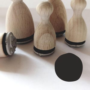 Può includere: Cinque timbri in legno con cuscinetti d'inchiostro neri. I timbri hanno la forma di un birilli da bowling e hanno una base rotonda. I timbri sono disposti a semicerchio su una superficie bianca.