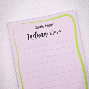 Könnte beinhalten: Ein hellvioletter Notizblock mit einem lindgrünen Rand und horizontalen Linien zum Schreiben. Oben auf der Seite steht der durchgestrichene Text "To do Liste", gefolgt von dem fettgedruckten, schwarzen Text "Tadaaa Liste" darunter.