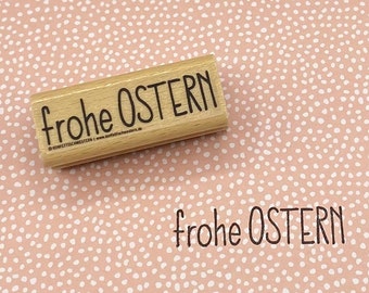 Può includere: Un timbro in legno con la scritta "frohe OSTERN" in nero, insieme a un'impressione dello stesso testo. Il timbro è rettangolare e il testo è in stile scritto a mano. Lo sfondo è una superficie rosa con pois bianchi.