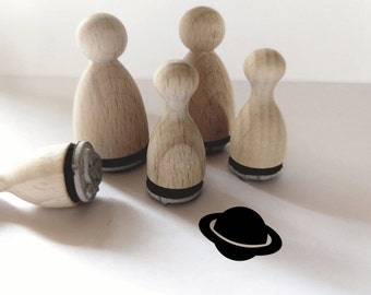 May include: Four wooden stamps with black ink pads, shaped like people, and a fifth stamp shaped like a planet. The planet stamp is black and has a ring around it. The stamps are on a white surface.