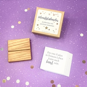 May include: A brown cardboard box with a white label that says "Adventskalender 24 K&auml;rtchen mit positiven Gedanken". The box is sitting on a purple surface with gold and silver confetti. A wooden stand and a stack of white cards with black text that says "Der rote Faden in Deinem Leben kann auch bunt sein." are also on the surface.