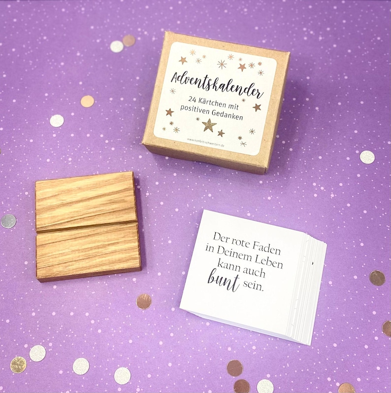 May include: A brown cardboard box with a white label that says "Adventskalender 24 K&auml;rtchen mit positiven Gedanken". The box is sitting on a purple surface with gold and silver confetti. A wooden stand and a stack of white cards with black text that says "Der rote Faden in Deinem Leben kann auch bunt sein." are also on the surface.