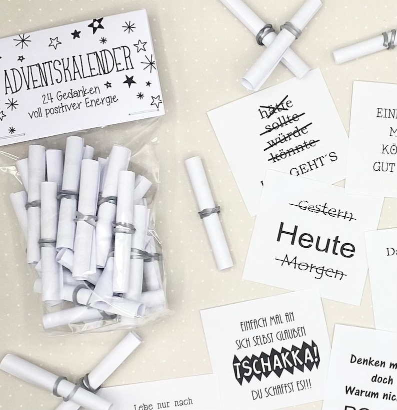 May include: A white bag filled with rolled up pieces of paper with black text that reads "Adventkalender 24 Gedanken voll positiver Energie."  The bag is on a white surface with several white pieces of paper with black text.  The text on the paper reads "hatte sollte w&uuml;rde k&ouml;nnte GEHT'S -Gestern Heute -Morgen EINFACH MAL AN SICH SELBST GLAUBEN TSCHAKKA! DU SCHAFFST ES!!! Denke doch Warum nic."