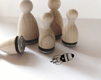 May include: Five wooden stamps with black ink pads, shaped like people, and a rocket stamp with black ink. The rocket stamp is on a white surface.