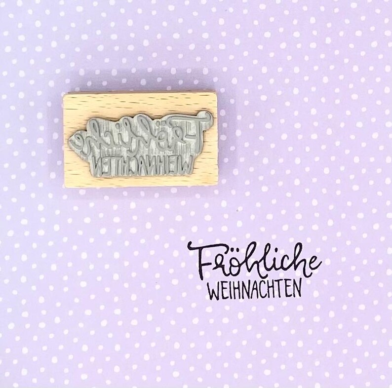 Peut inclure: Un tampon en bois avec le texte "Frohe Weihnachten" en &eacute;criture cursive. Le tampon est sur un fond violet avec des pois blancs.
