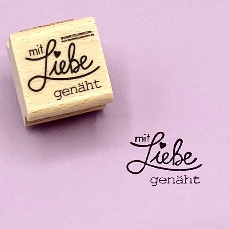 May include: Wooden stamp with the text "mit Liebe gen&auml;ht" which translates to "with love sewn" in English. The stamp has a small heart symbol above the word "Liebe".