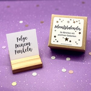 May include: A small wooden box with a brown paper label that says "Adventskalender 24 K&auml;rtchen mit positiven Gedanken" and a gold star design. The box is sitting on a purple surface with silver and pink confetti. A small wooden stand holds a white card with the text "Folge deinem Funkeln."