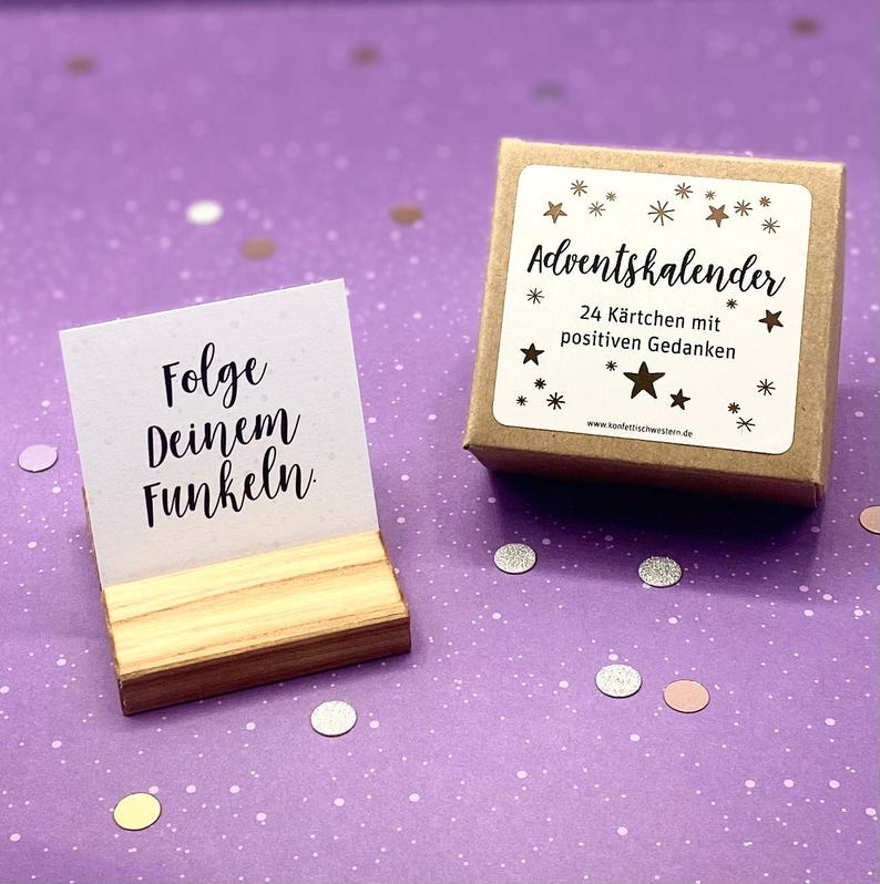 May include: A small wooden box with a brown paper label that says "Adventskalender 24 K&auml;rtchen mit positiven Gedanken" and a gold star design. The box is sitting on a purple surface with silver and pink confetti. A small wooden stand holds a white card with the text "Folge deinem Funkeln."