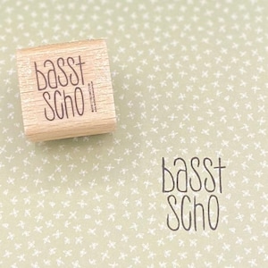 Op de afbeelding: Een houten stempel met de tekst "basst scho" op een groene achtergrond met een wit patroon.