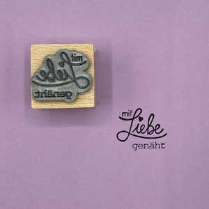 May include: A wooden stamp with the text "gen&auml;ht mit Liebe" in a cursive font. The stamp is on a light purple background.