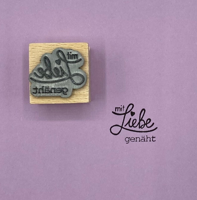 May include: A wooden stamp with the text "gen&auml;ht mit Liebe" in a cursive font. The stamp is on a light purple background.