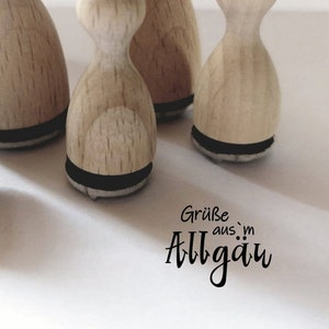 Può includere: Quattro timbri in legno con cuscinetti d'inchiostro neri. I timbri hanno la forma di un rettangolo arrotondato con un piccolo manico in cima. I timbri sono disposti in fila su una superficie bianca. Il testo "Grüße aus dem Allgäu" è stampato sulla superficie sotto i timbri.