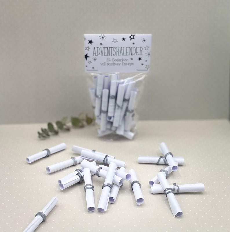 May include: A white plastic bag filled with rolled up white paper slips tied with gray rubber bands. The bag has a label that reads "Adventskalender 24 Gödanken voll positiver Energie".