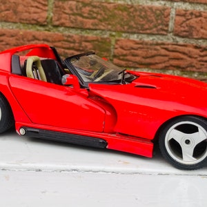 Puede incluir: Un coche modelo Dodge Viper rojo con interior negro y ruedas plateadas. El coche de juguete es un descapotable con la capota bajada, y la palabra "Viper" es visible en el lateral.