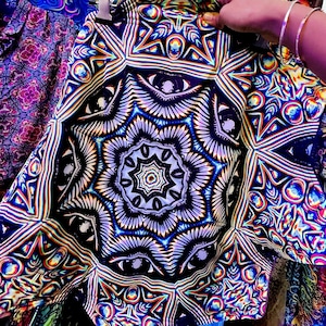 May include: A patterned garment with a geometric design, featuring eye-like shapes and a central starburst. The fabric displays a vibrant mix of blues, yellows, and purples, creating a psychedelic effect. Other similar garments are visible in the background.