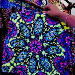 Puede incluir: Un textil vibrante con un diseño de caleidoscopio en tonos azules, rosas y verdes. La tela está colgada de una percha metálica. El tema general es el arte psicodélico.