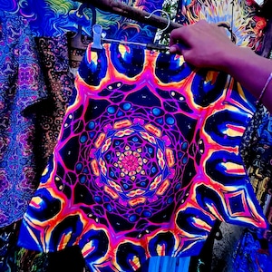 Può includere: Un capo dai vivaci motivi psichedelici, forse pantaloncini o una gonna, è appeso a un gancio. Il design presenta un mandala centrale con motivi radiali nei toni del blu, viola, arancione e giallo. Altri tessuti con motivi simili sono visibili.