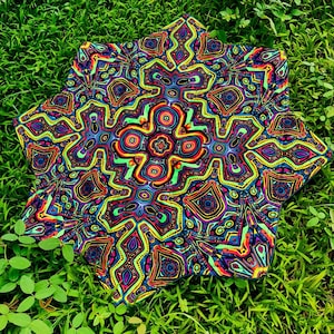 Puede incluir: Textil en forma de estrella con un patrón psicodélico. El diseño presenta una compleja disposición de formas coloridas en tonos de azul, verde, amarillo, naranja y morado. El textil está sobre un fondo de hierba verde.