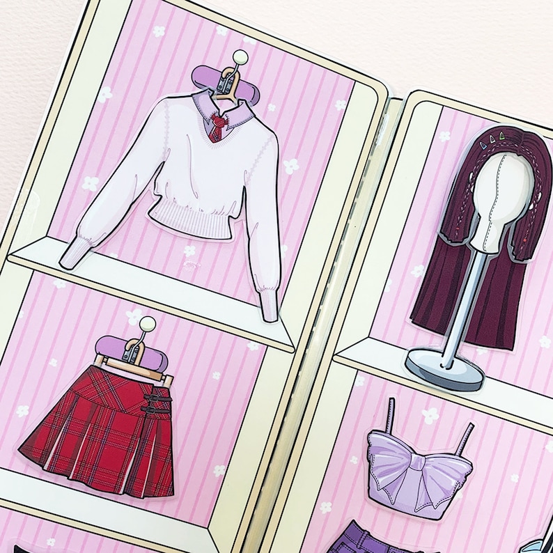 May include: A pink and white closet with three shelves. The top shelf has a white sweater with a collar and a bow tie. The middle shelf has a red and black plaid skirt. The bottom shelf has a purple top with a bow. The closet is decorated with small white flowers.