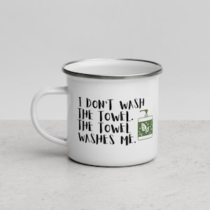 May include: White enamel mug with a black and green design. The mug has a silver rim and handle. The text on the mug reads "I don't wash the towel. The towel washes me."