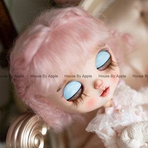 May include: A pink haired doll with closed eyes and blue eye shadow. The doll is wearing a white dress with lace trim.