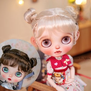 May include: Two dolls with different hairstyles. The doll on the left has brown hair with bangs and two buns. The doll on the right has blonde hair with two buns. Both dolls have large, blue eyes and are wearing dresses.