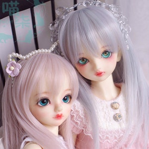May include: Two dolls with long, flowing hair. The doll on the left has pink hair and is wearing a white dress. The doll on the right has silver hair and is wearing a pink dress with white lace. Both dolls have blue eyes and are holding a pink bunny.