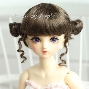 May include: A brown wig with curly bangs and two buns on top. The wig is styled on a doll with blue eyes and a pink dress.