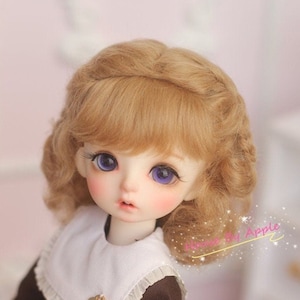 May include: A doll with blonde curly hair, wearing a brown dress with a white collar and a small gold button. The doll has blue eyes and a light pink complexion.