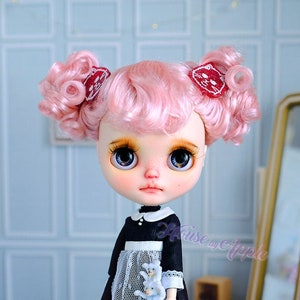 May include: A Blythe doll with pink curly hair and red bows. The doll wears a black dress with a white lace apron and red shoes. She holds a small blue stuffed animal. A small doll with white fur is in the foreground.