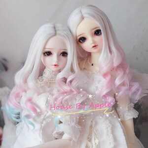May include: Two dolls with long, white wigs with pink and blue ombre highlights. The wigs are styled in loose curls. The dolls are wearing white dresses with lace trim. The text "House By Apple" is visible in the image.