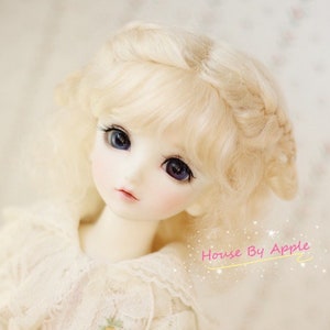 May include: A doll with blonde curly hair styled in a braid. The doll has blue eyes and is wearing a white dress with lace trim. The doll is looking to the side and has a soft, innocent expression.