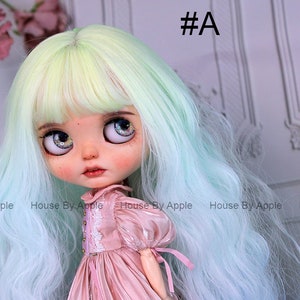 May include: A doll with long, wavy, light green and blue hair, wearing a pink dress with lace details. The doll has large, blue eyes with glitter.