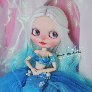 May include: A doll with long, white and blue hair, wearing a blue and white seashell bra and a blue sequined mermaid tail. The doll is sitting on a blue tulle skirt and has a pink background.
