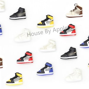 Könnte beinhalten: Eine Sammlung von Miniatur-High-Top-Sneakern in verschiedenen Farben, darunter Schwarz, Weiß, Gelb, Rot und Blau. Jeder kleine Schuh verfügt über Schnürsenkel, ein Seitenteil und eine Gummisohle. Der Text House By Apple ist in der Mitte sichtbar.