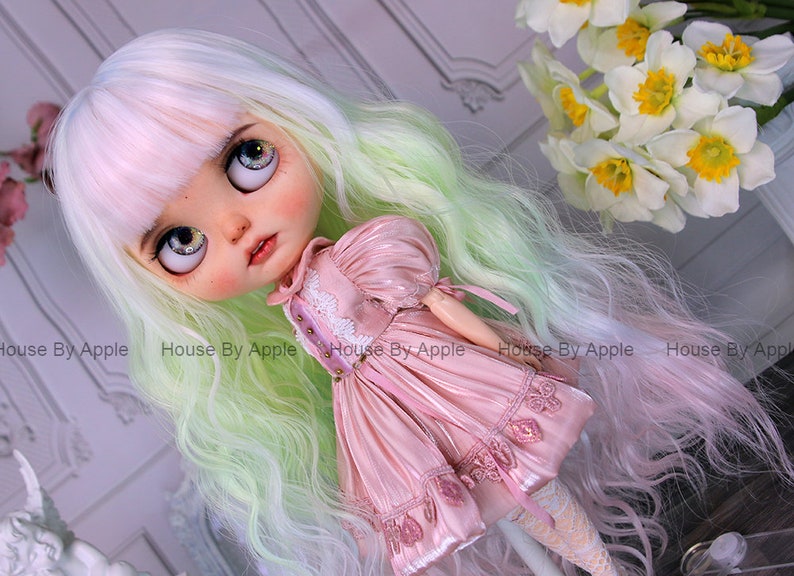 May include: A doll with long, wavy, green and pink hair, wearing a pink dress with lace trim. The doll has large, blue eyes and a painted face.
