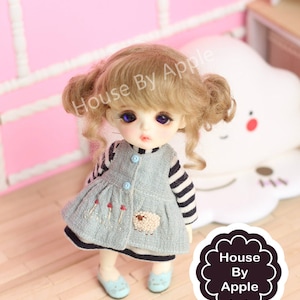 May include: A small doll with blonde curly hair, blue eyes, and a light blue dress with a sheep embroidered on the front. The doll is wearing a striped shirt underneath the dress and has blue shoes. The doll is standing on a wooden floor.