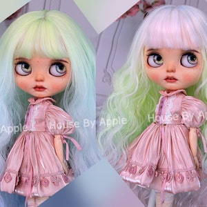May include: Two dolls with long, wavy hair in different colors. The doll on the left has light green and blue hair, while the doll on the right has white and pink hair. Both dolls are wearing a pink dress with lace details and are holding a small white flower.
