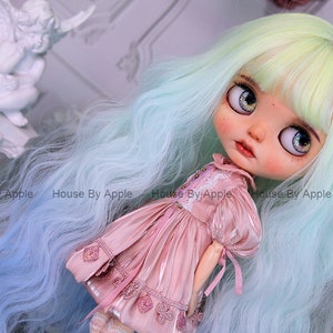 May include: A doll with long, wavy, light blue and green hair, wearing a pink dress with lace details. The doll has large, blue eyes with glitter and a painted face.