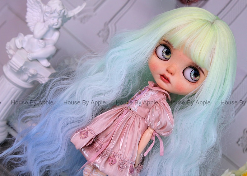 May include: A doll with long, wavy, light blue and green hair, wearing a pink dress with lace details. The doll has large, blue eyes with glitter and a painted face.