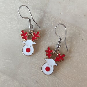 Può includere: Un paio di orecchini a forma di renna natalizia. Ogni orecchino presenta una testa di renna bianca con naso e corna rosse, bordata d'oro. Gli orecchini hanno ganci per orecchie color argento, perfetti per le feste.