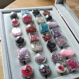 Peut inclure: Une collection de bagues faites à la main exposées dans un écrin gris. Les bagues présentent des dômes en résine transparente remplis de paillettes, de perles et d'éléments floraux de différentes couleurs, dont le rose, le rouge, le noir et l'argent. Les bagues sont sur des bagues en argent.