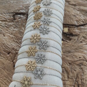 Peut inclure: Présentation de bracelets à breloques flocon de neige en or et argent. Les bracelets sont disposés sur un coussin en tissu blanc avec la marque "Lolilota" et le mot "Acier" imprimés. Le fond est une surface en bois.