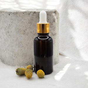 May include: A dark amber glass bottle with a gold-colored collar and white dropper. The bottle is next to several small, yellow dried flowers. The background includes a concrete block and a white cloth.