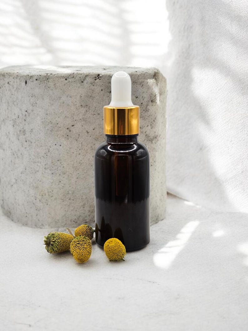 May include: A dark amber glass bottle with a gold-colored collar and white dropper. The bottle is next to several small, yellow dried flowers. The background includes a concrete block and a white cloth.