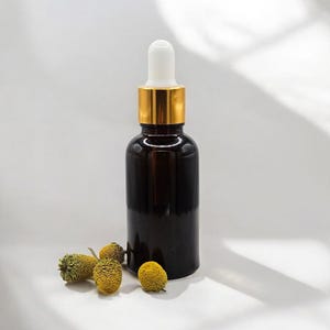 May include: A dark amber glass dropper bottle with a gold collar and white dropper top. Several dried yellow flowers are scattered near the base of the bottle. The bottle is likely for essential oils or serums.