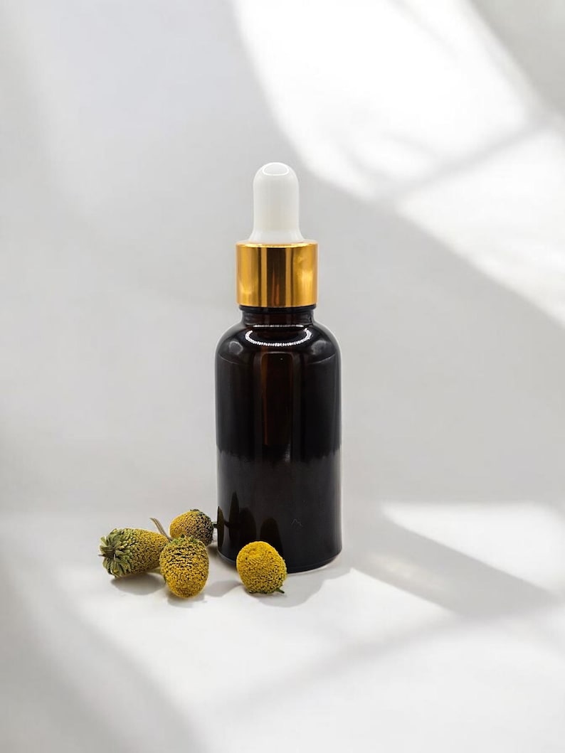 May include: A dark amber glass dropper bottle with a gold collar and white dropper top. Several dried yellow flowers are scattered near the base of the bottle. The bottle is likely for essential oils or serums.