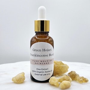 May include: A brown glass bottle with a gold lid and a white dropper. The bottle is labeled "Green Hojari Frankincense Resin Pure Nourish Skincare Oleo Extract 100% Organic Jojoba Oil External use only". There are several pieces of frankincense resin on a white surface.