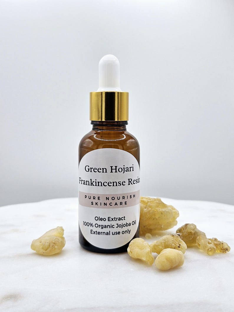 May include: A brown glass bottle with a gold lid and a white dropper. The bottle is labeled "Green Hojari Frankincense Resin Pure Nourish Skincare Oleo Extract 100% Organic Jojoba Oil External use only". There are several pieces of frankincense resin on a white surface.